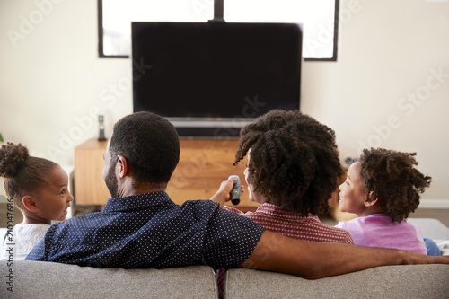Rear View Of Family With Children Sitting On Sofa Watching TV Together