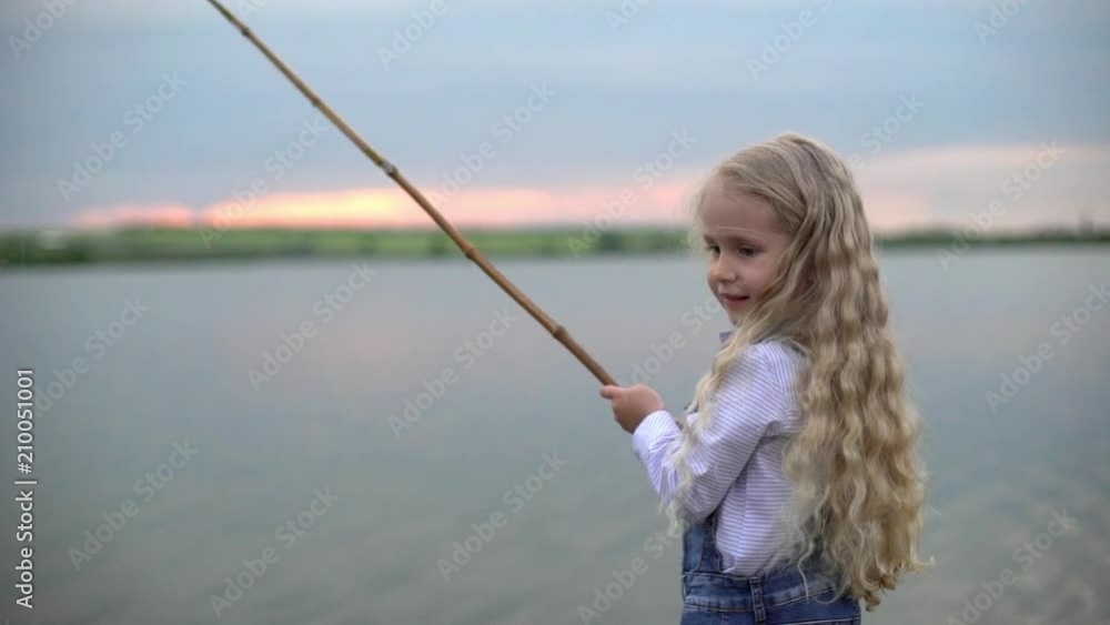 Portrait slow motion closeup of beautiful european girl 7-8 with blonde long hair fishing on wooden pier and putting rod in water while spending weekend on lake or river during sunset Portrait slow motion closeup of beautiful european girl 7-8 with blonde long hair fishing on wooden pier and putting rod in water while spending weekend on lake or river during sunset