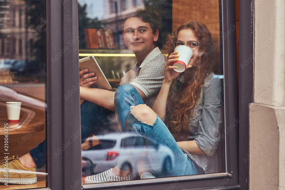 Couple of young students drinking coffee and using a digital tablet while sitting on a window sill at a college campus during a break.