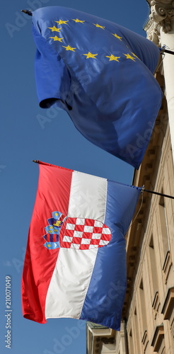 Flagi Chorwacji i Unii Europejskiej powiewają na wietrze, zawieszone obok sie, na zenętrznej ścianie budynku, w tle niebieskie niebo