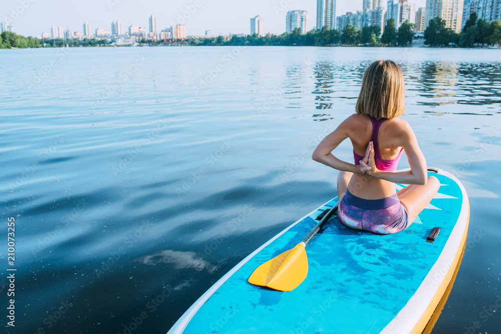 Young woman practicing yoga on a paddleboard In city bay, back view