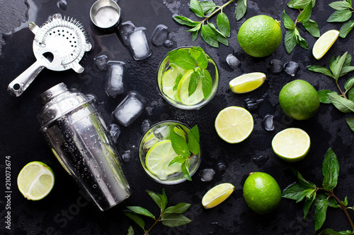 Mojito cocktail alcohol bar long drink making. Mint, lime, rum, ice ingredients, juice, soda water, shaker and bar utensils on black stone background. Top view. Flat lay.