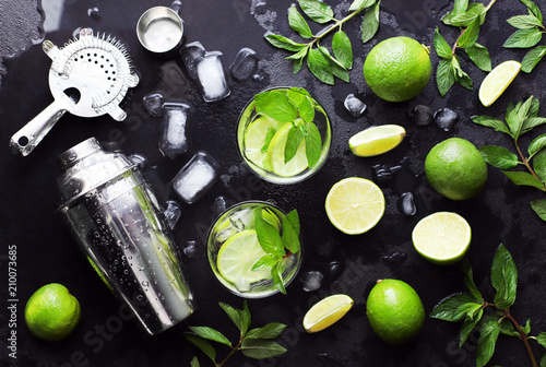 Mojito cocktail Cuba tropical alcohol bar long drink, two highball glass with rum, mint, lime, shaker, juice, soda water and ice on black stone background. Flat lay. Top view.