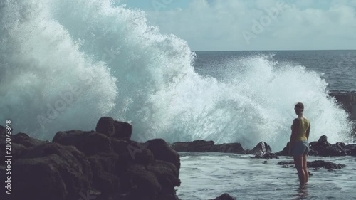 SLOW MOTION: Young female traveler watching the big waves violently crash into the rocky shore. Foaming ocean water splashes high into the air and over the black rocks of a coast on Easter Island.