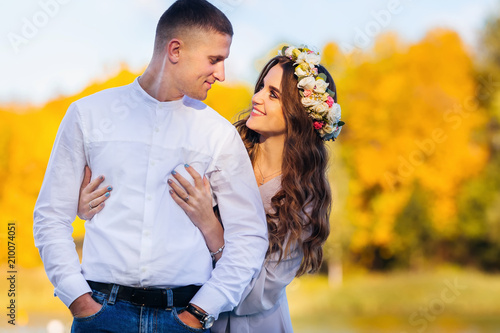 A pretty man holds his hands in his pockets and his wife hugs him against the background of yellow trees