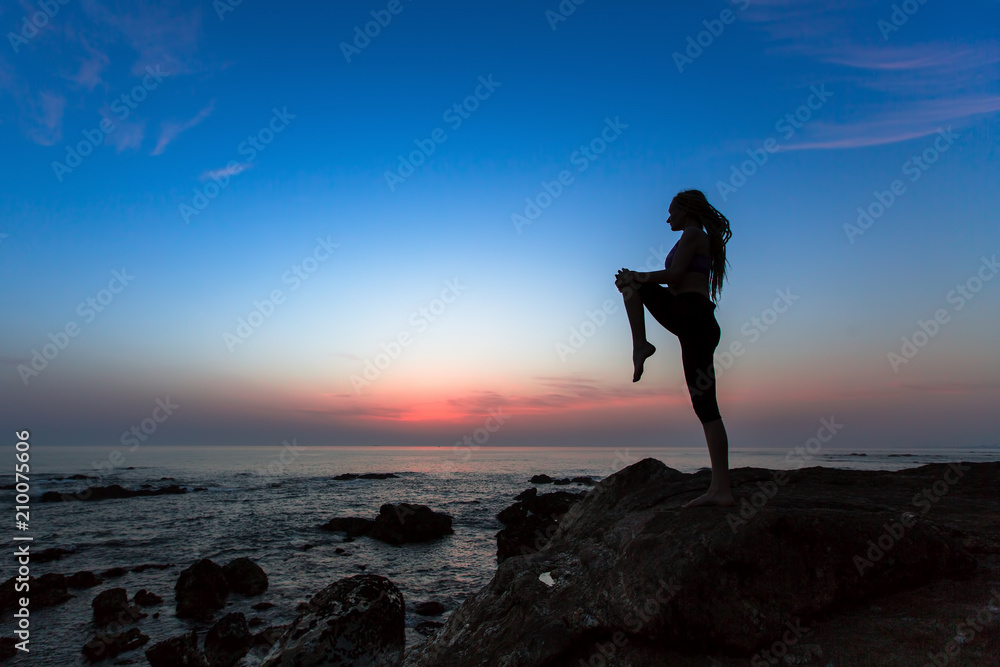 Silhouette of a young woman practicing yoga exercises on the ocean at twilight.