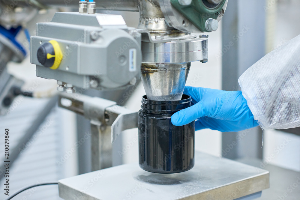 Crop close-up production worker hand in protective glove filling black container with sports nutrition. Crop close-up production worker hand in protective glove filling black container with sports nutrition.