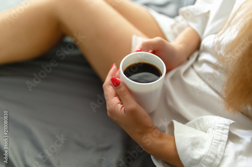 Slim, perfect and beautiful woman drinking her coffee while sitting on bed. Sunny Lazy morning in bed. Cropped image of erotic lying on the bed of a girl in the bedroom. Top view.