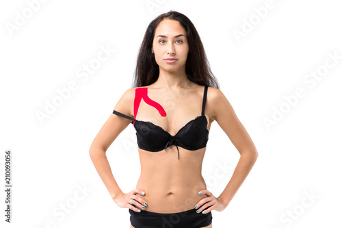 The theme kinesiology taping, help stretching, trauma and muscle pain. Young caucasian woman with long brunette hair posing in black lingerie with red lipstick tape on shoulder on isolated background
