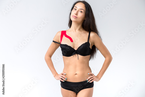 The theme kinesiology taping, help stretching, trauma and muscle pain. Young caucasian woman with long brunette hair posing in black lingerie with red lipstick tape on shoulder on isolated background
