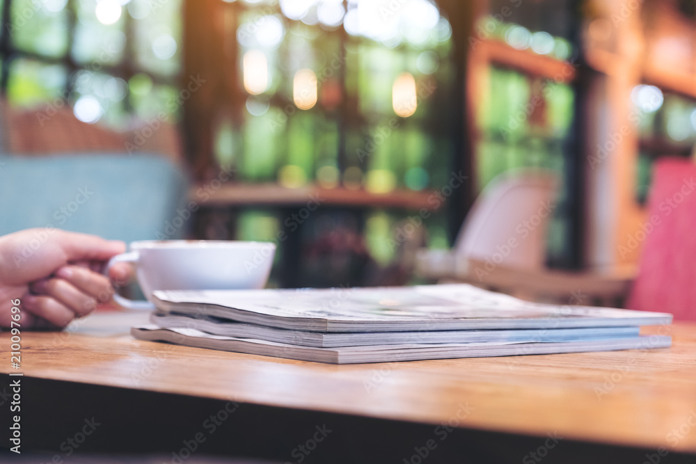 Closeup image of a hand holding white cup of hot coffee with the books on wooden table in cafe