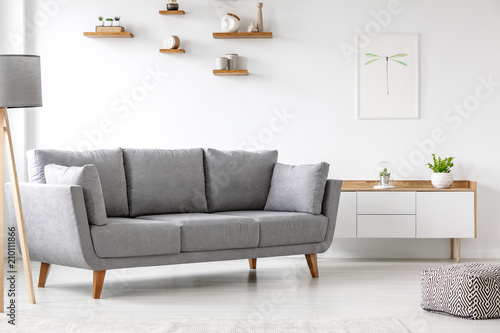 Simple, gray sofa standing next to a white cupboard in living room interior with decorations on wooden shelves. Real photo photo