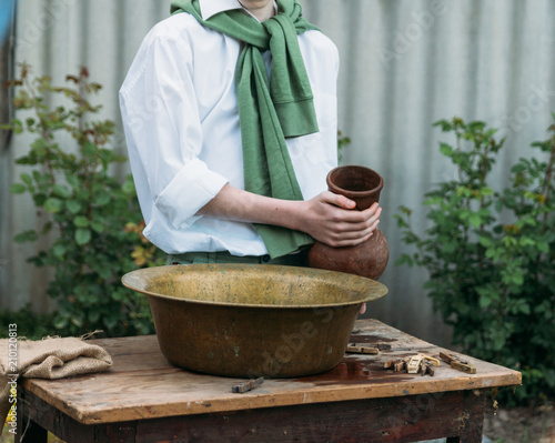 the boy holds a clay jug, pours water out of it into a copper basin. help photo