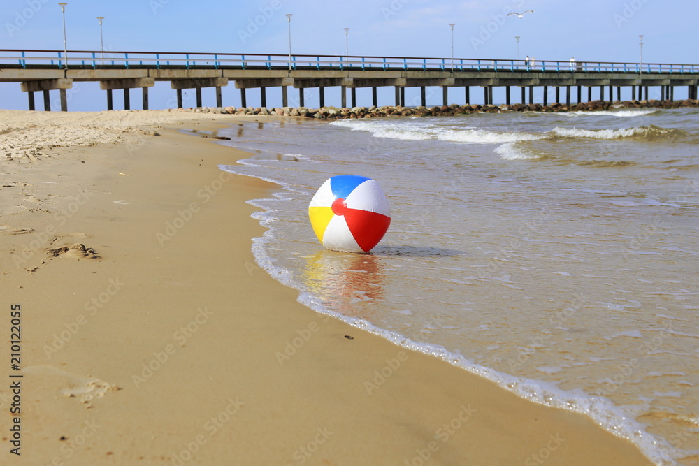 Obraz premium Colorful beach ball on sand and waving sea. Holiday beach ball near Baltic sea and Palanga bridge in horizon.