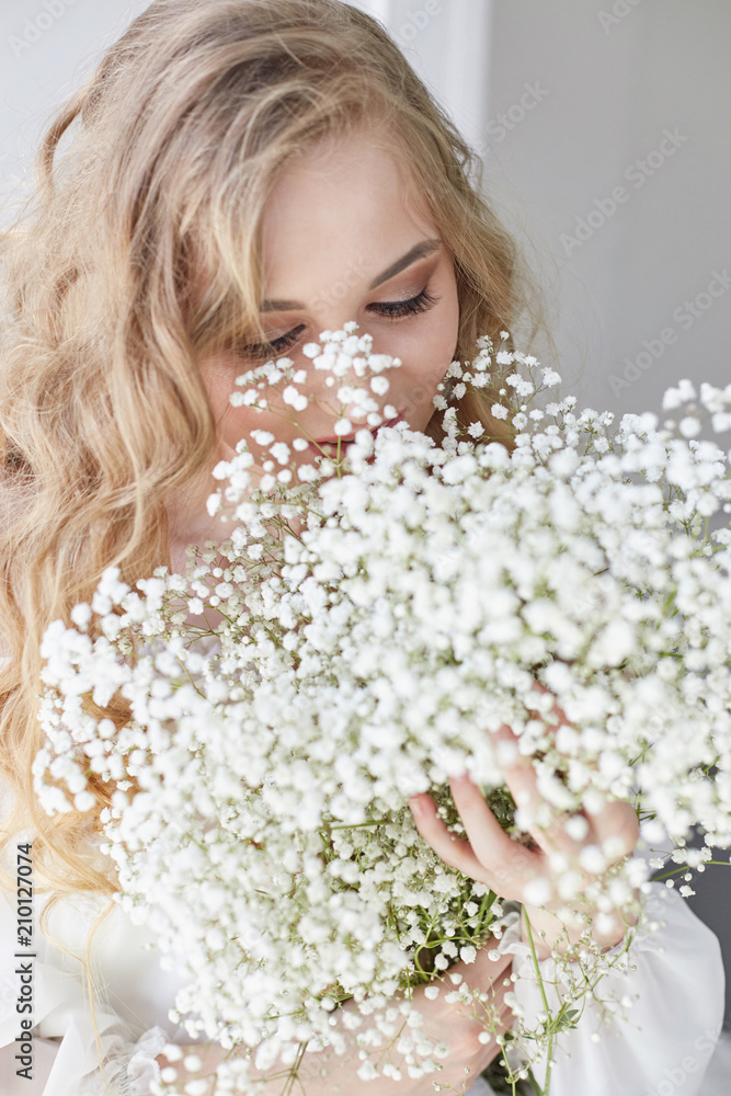 Naklejka premium Curly blonde romantic look, beautiful eyes. White wildflowers in hands. Girl white light dress and curly hair, portrait of woman with flowers at home near the window, purity and innocence