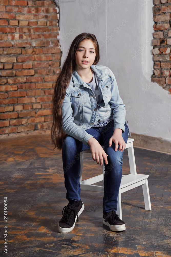 Beautiful stylish hipster girl sitting chair,looking camera. Attractive teenager fashion pose. Adolescent woman wears casual fashionable clothes.Youth trend.Studio shot, loft interior, style.