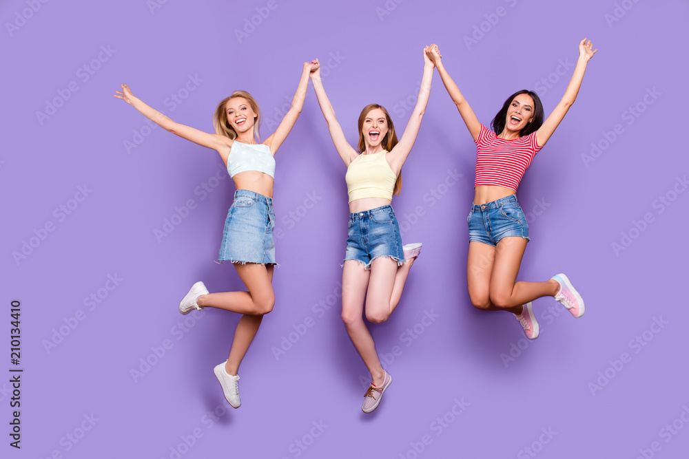 Portrait of crazy funky girls in jeans shorts skirt jumping in air holding raised arms celebrating achievement yelling looking at camera isolated on bright violet background