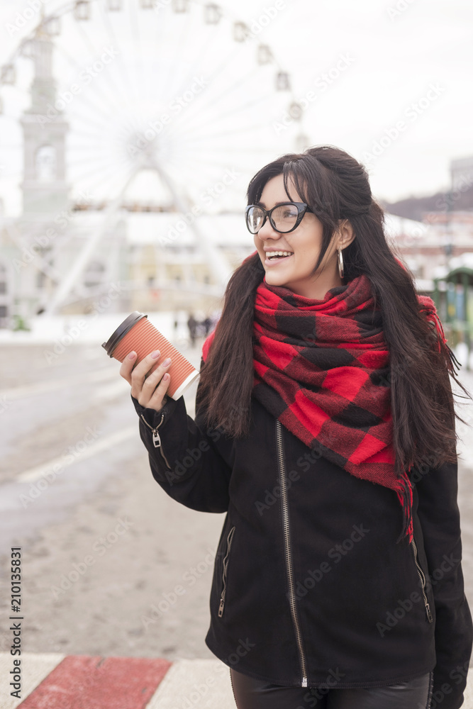 Cute young caucasian brunette woman student on a walk in europe city streets. She wearing blac coat and plaid red scarf. Cold winter weather. Woman drinks coffee from a to-go cup