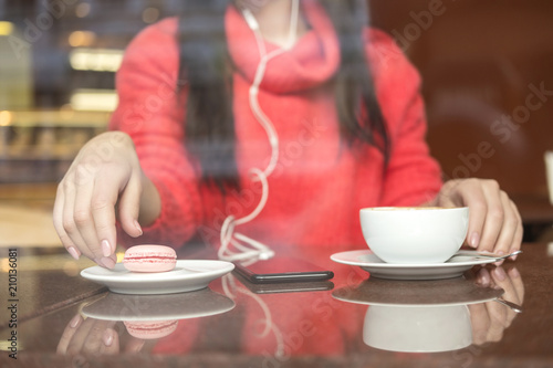 Cute brunette plus size woman working on a distance  freelance   sitting in cafe  drinking coffee and eating macaron. She wearing red sweater  listen music and typing messages on her phone