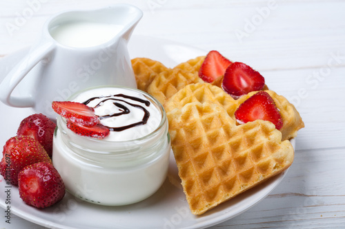 Heart shaped waffles with strawberries, cream sauce, pitcher with milk on white plate and white wooden background. St Valentines day breakfast. Romantic breakfast dessert. Copy space