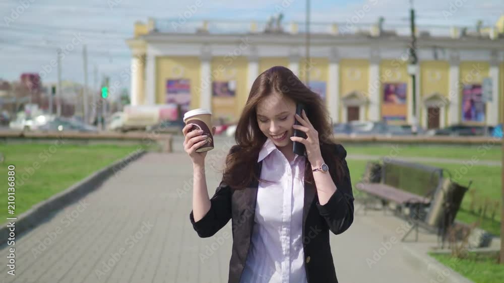 Beautiful brunette woman walking the city street and talking on the cell phone. Woman walking with coffee to go. Beautiful brunette woman walking the city street and talking on the cell phone. Woman walking with coffee to go.