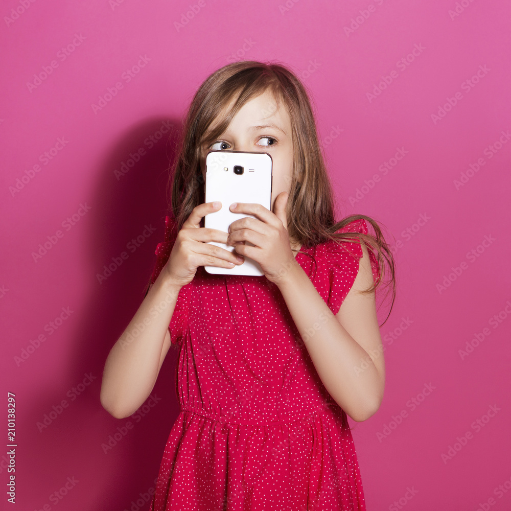 Little 8 years old girl make some emotional gesture with her hands on a pink neutral background. She has long brunette hair and wear red summer dress. Funny expression on her face