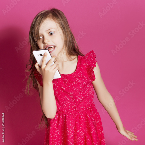 Little 8 years old girl make some emotional gesture with her hands on a pink neutral background. She has long brunette hair and wear red summer dress. Funny expression on her face