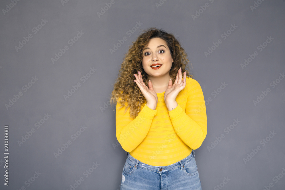 Cute brunette plus size woman with curly hair in yellow sweater and jeans standing on a neutral grey background. She amazed, shocked about some surprise Cute brunette plus size woman with curly hair in yellow sweater and jeans standing on a neutral grey background. She amazed, shocked about some surprise