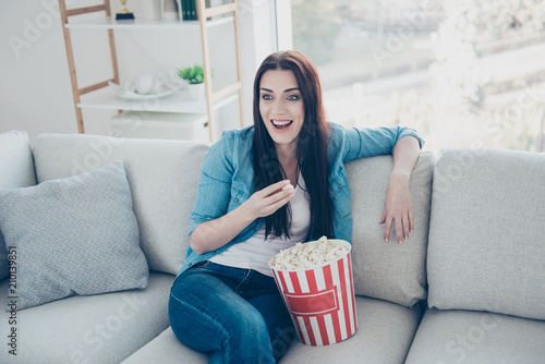 Portrait of positive toothy woman in jeans outfit watching comedy having bucket of pop-corn laughing enjoying funny film sitting in livingroom