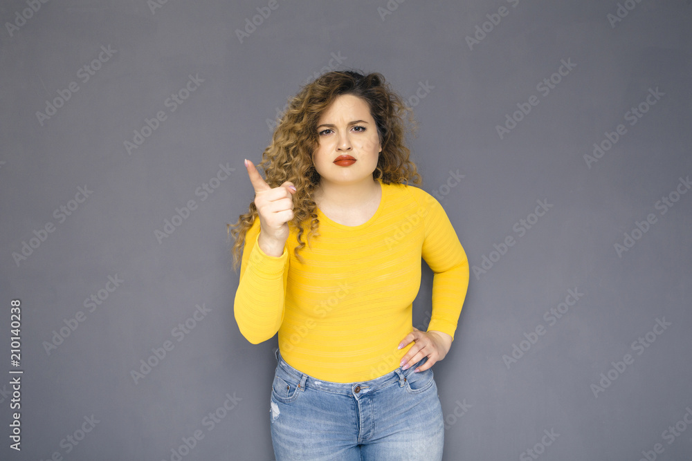 Cute brunette plus size woman with curly hair in yellow sweater and jeans standing on a neutral grey background. She threat someone, making gesture using her hand Cute brunette plus size woman with curly hair in yellow sweater and jeans standing on a neutral grey background. She threat someone, making gesture using her hand