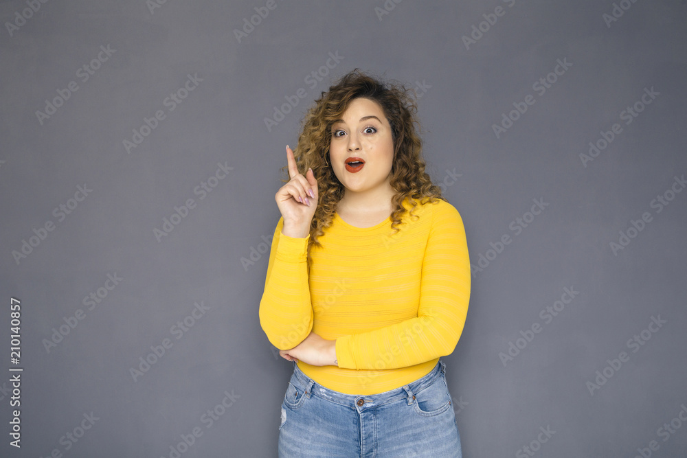 Cute brunette plus size woman with curly hair in yellow sweater and jeans standing on a neutral grey background. She was thinking, has an idea, thoughtful emotion on her face. Copy space Cute brunette plus size woman with curly hair in yellow sweater and jeans standing on a neutral grey background. She was thinking, has an idea, thoughtful emotion on her face. Copy space