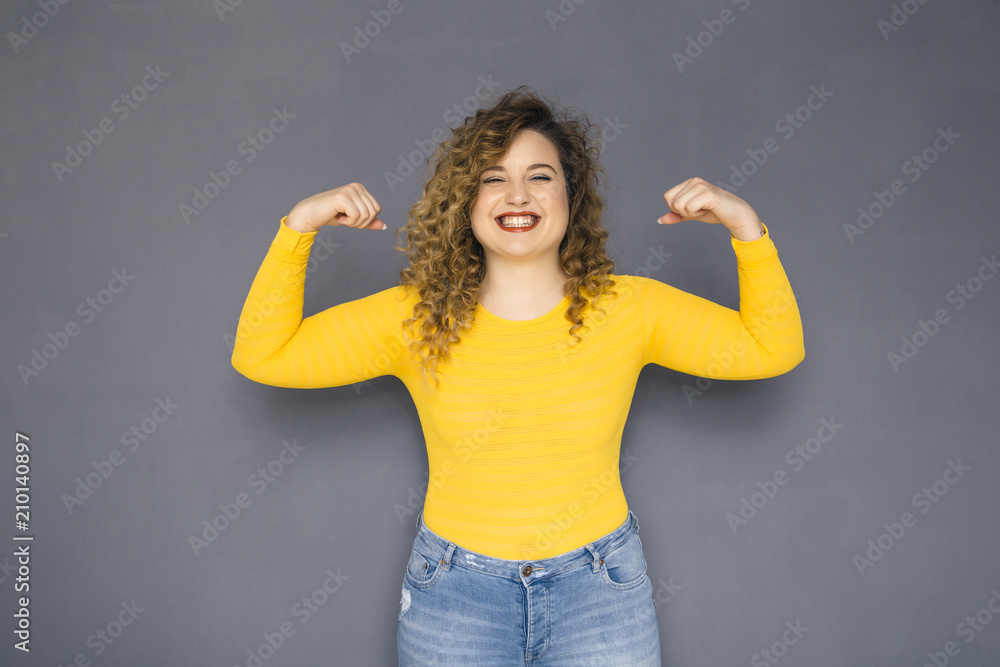 Cute brunette plus size woman with curly hair in yellow sweater and jeans standing on a neutral grey background. She shows her biceps. Powerful pose Cute brunette plus size woman with curly hair in yellow sweater and jeans standing on a neutral grey background. She shows her biceps. Powerful pose