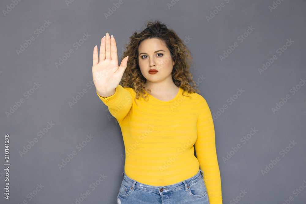 Cute brunette plus size woman with curly hair in yellow sweater and jeans standing on a neutral grey background. She makes a stop sign using her hand Cute brunette plus size woman with curly hair in yellow sweater and jeans standing on a neutral grey background. She makes a stop sign using her hand