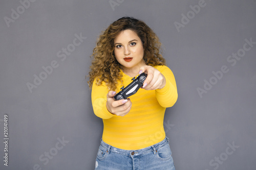 Cute brunette plus size woman with curly hair in yellow sweater and jeans standing on a neutral grey background. She holds a joystick, playing on console games
