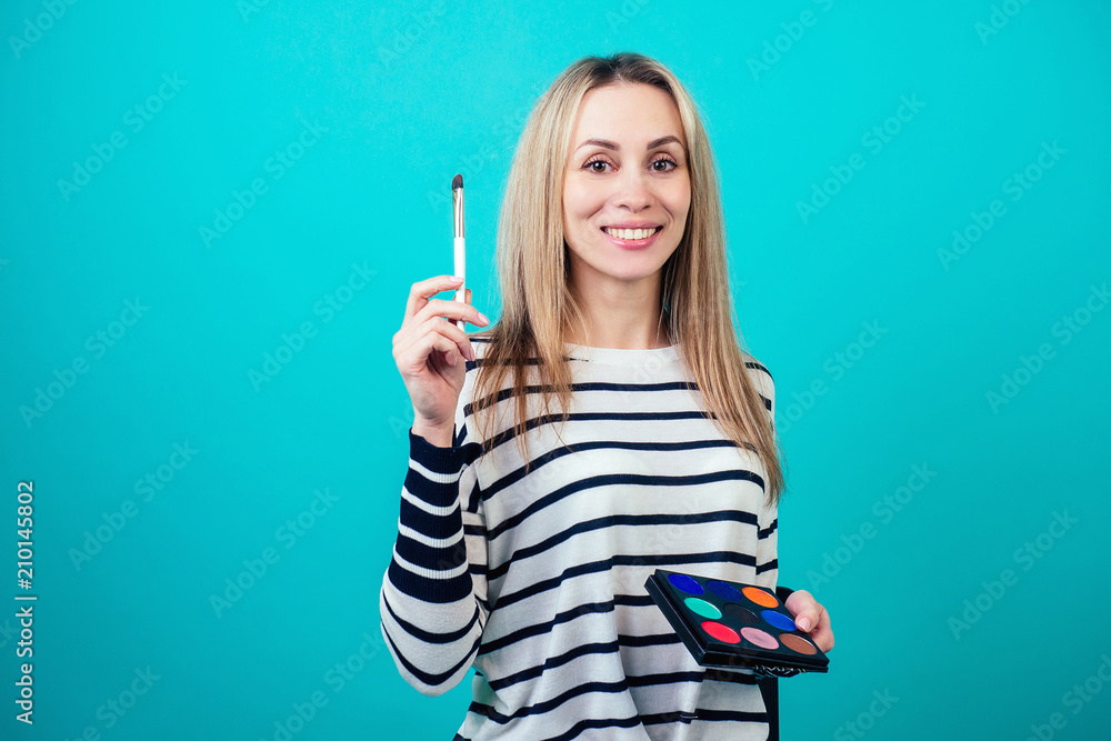Obraz premium young and attractive blonde woman visagiste (make-up artist ) holds a brush makeup on blue background in a studio. concept of skin care and beauty cosmetics for face