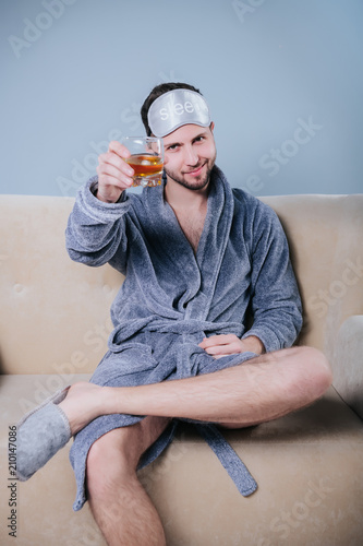 A serious guy with a beard holds a glass of cola or whiskey with ice in his hand.Young playboy in dressing gown toasting with cocktail.Happy young man in dressing gown drinking martini