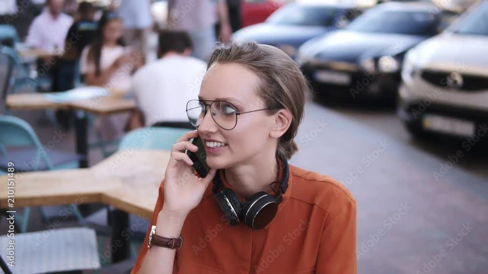 Close up of fair hair girl with headphones on her neck talking on mobile phone while sitting in street cafe. Smiling, wearing stylish glasses, dressed in orange blouse Close up of fair hair girl with headphones on her neck talking on mobile phone while sitting in street cafe. Smiling, wearing stylish glasses, dressed in orange blouse