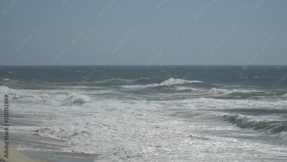 View of a beautiful ocean sandy beach with the blue waves rolling into the shore, some rocks present. View of a beautiful ocean sandy beach with the blue waves rolling into the shore, some rocks present.