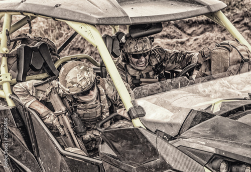 Armed modern infantry soldiers, elite members of special forces, private military company mercenaries extremely driving desert patrol vehicle while going at rough terrain. Desaturated, cropped shoot photo