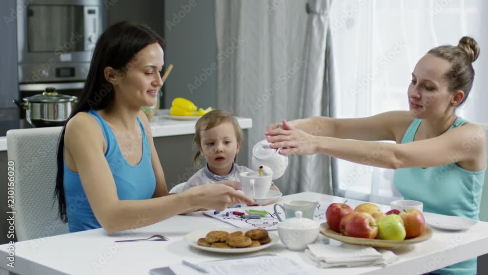 PAN of cheerful young woman pouring hot tea into cup of female partner while having breakfast in morning, then looking at cute toddler girl coloring with crayons PAN of cheerful young woman pouring hot tea into cup of female partner while having breakfast in morning, then looking at cute toddler girl coloring with crayons