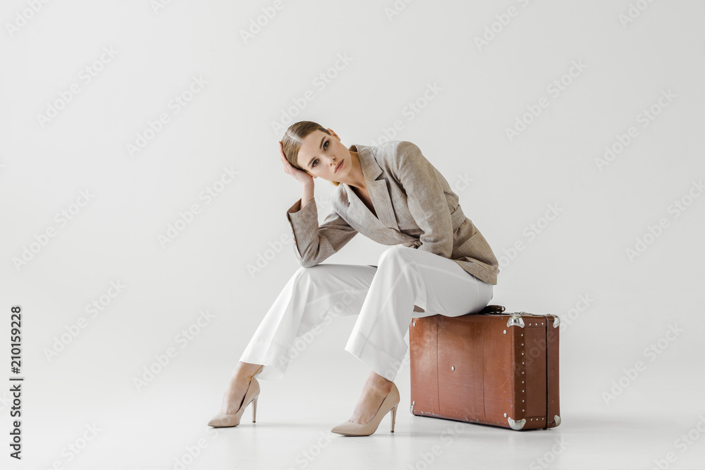young stylish female model in linen jacket sitting on vintage suitcase and looking at camera isolated on grey background - obrazy, fototapety, plakaty 