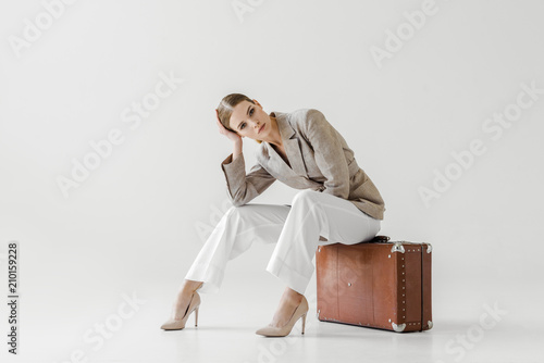 young stylish female model in linen jacket sitting on vintage suitcase and looking at camera isolated on grey background