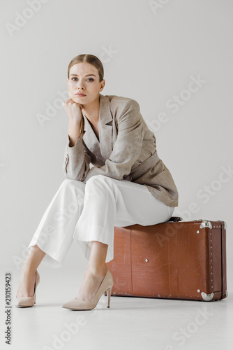 stylish young female model in linen jacket with hand on chin sitting on vintage suitcase isolated on grey background
