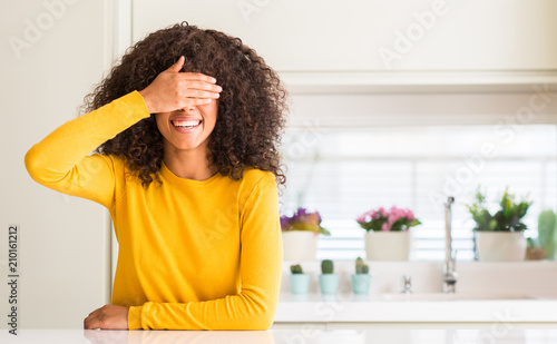 African american woman wearing yellow sweater at kitchen smiling and laughing with hand on face covering eyes for surprise. Blind concept.