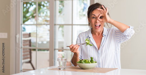 Middle aged woman eating fresh salad in a bowl at home with happy face smiling doing ok sign with hand on eye looking through fingers