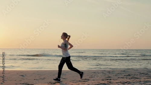 Beautiful Slender Woman is Running along Sea Coast at Sunset and Jogging on Beach with Headphones in SlowMotion