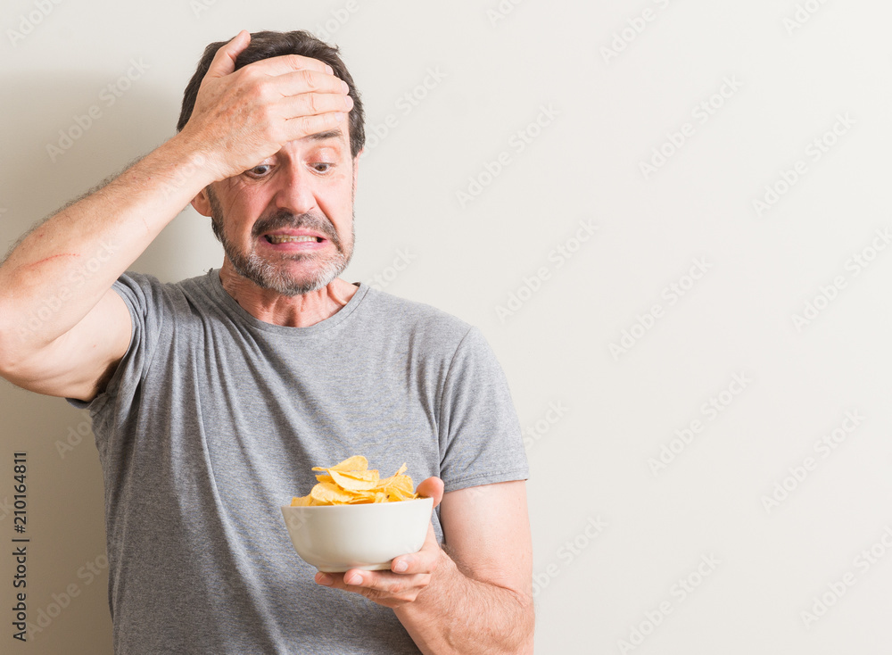 Senior man eating potato chips stressed with hand on head, shocked with