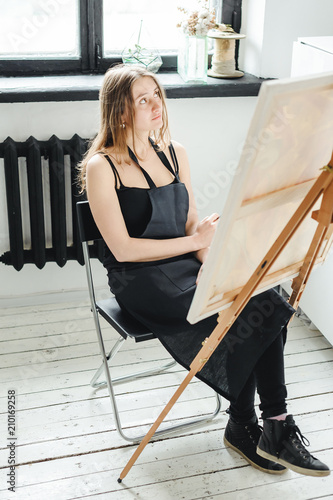 young woman in black clothes and an apron sits in a bright room and paints pictures, brushing oil paints on canvas with malbert photo