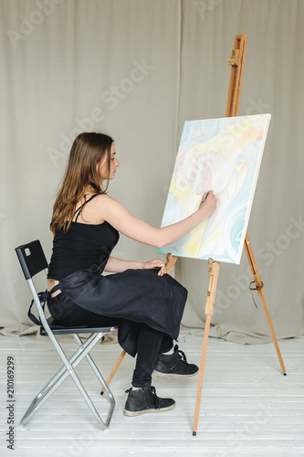 young woman in black clothes and an apron sits in a bright room and paints pictures, brushing oil paints on canvas with malbert photo