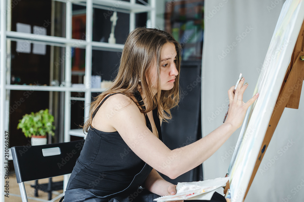 Obraz premium young woman in black clothes and an apron sits in a bright room and paints pictures, brushing oil paints on canvas with malbert